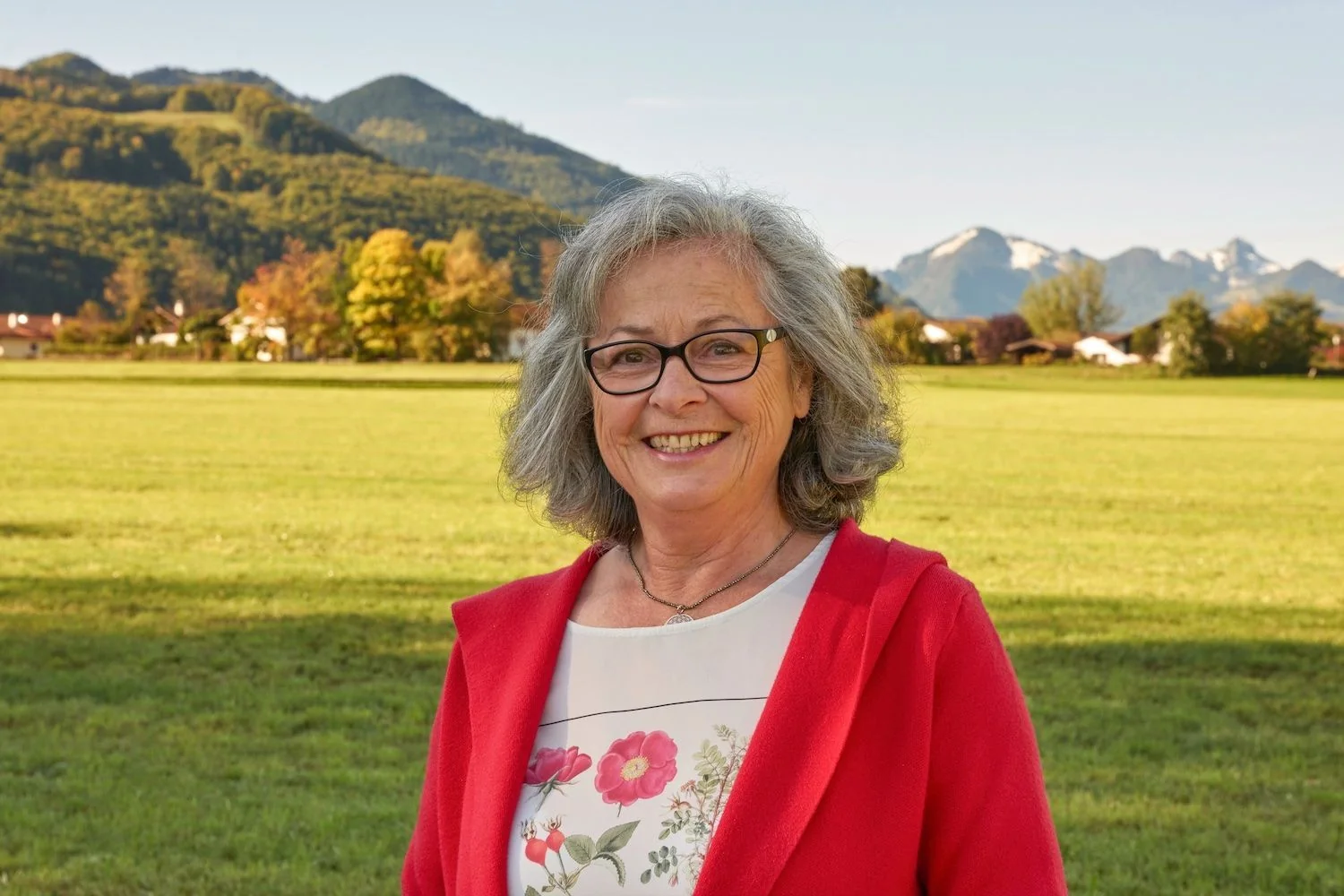 Irmengard Linder, Kräuterpädagogin am Chiemsee, im roten Cardigan vor Wiesen und Chiemgauer Bergen